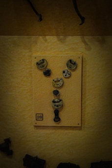 A collection of aged metal locks and keys displayed on a board with a numbered label '10'. The board is mounted on a textured yellow wall, and there are shadows implying a dim lighting setting.