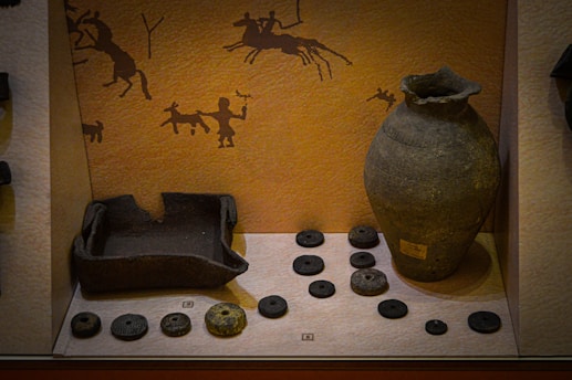 The image depicts a museum display featuring ancient pottery and artifacts. A large, intricately designed clay vase stands prominently on the right. To the left, several circular stone objects are arranged, likely serving as weights or ornaments. A broken pot is also visible. The back wall is decorated with traditional cave art-style drawings, including figures of humans and animals, suggesting a historical or cultural context.