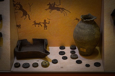 The image depicts a museum display featuring ancient pottery and artifacts. A large, intricately designed clay vase stands prominently on the right. To the left, several circular stone objects are arranged, likely serving as weights or ornaments. A broken pot is also visible. The back wall is decorated with traditional cave art-style drawings, including figures of humans and animals, suggesting a historical or cultural context.