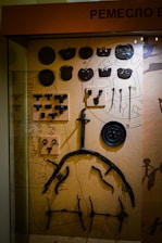 A display case containing various historical metal artifacts arranged on a textured background. The items include round and irregularly shaped pieces, hooks, and tools. There is a mix of symmetrical and asymmetrical objects, possibly related to historic trades or craftsmanship.