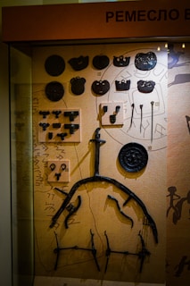 A display case containing various historical metal artifacts arranged on a textured background. The items include round and irregularly shaped pieces, hooks, and tools. There is a mix of symmetrical and asymmetrical objects, possibly related to historic trades or craftsmanship.