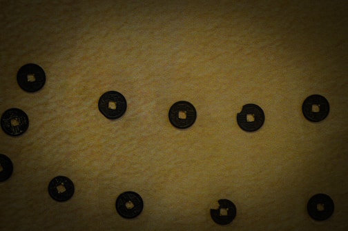 Several circular coins with square holes in the center are scattered on a textured brown surface. The coins appear dark with intricate details or inscriptions on them.