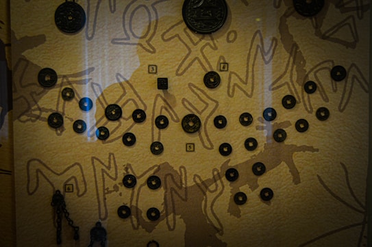 A collection of circular artifacts is displayed on a surface with a textured background featuring abstract patterns. The artifacts are arranged in a formation and numbered labels are attached to some of them, suggesting a categorization or order. The background has a warm, earthy tone with lighter lines creating a contrast.