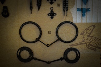 A display of several metallic objects, including a pair of large, circular handcuffs with intricate details and two other decorative metal pieces. The background appears to be a soft, beige textile with a subtle, printed pattern possibly resembling abstract shapes.