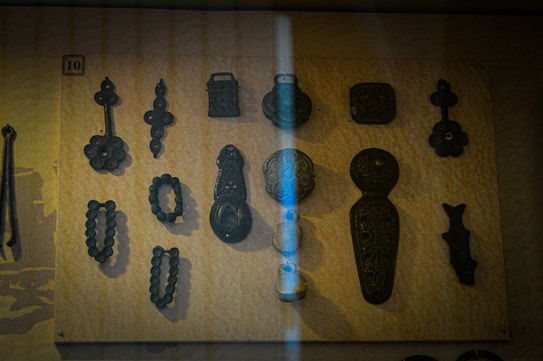 A collection of antique-looking metallic objects mounted on a board. The objects vary in shape and size, featuring intricate designs and appear to be historical artifacts. A vertical light stripe crosses the display, partly illuminating the objects.