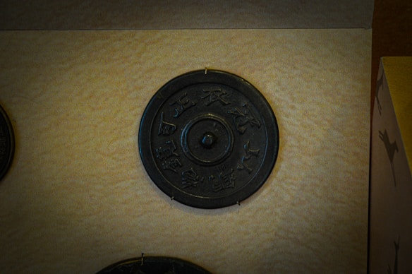 An ancient bronze mirror is displayed against a beige background. The circular mirror features intricate engravings and a raised central knob. Its historical and cultural significance is suggested by the ornate design.