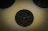 A round, dark metallic plaque with intricate Chinese characters embossed on its surface is mounted on a light beige textured wall. There are other plaques partially visible around it, each with unique designs.