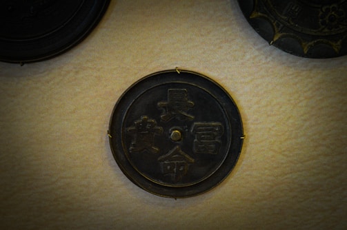 A round, dark metallic plaque with intricate Chinese characters embossed on its surface is mounted on a light beige textured wall. There are other plaques partially visible around it, each with unique designs.