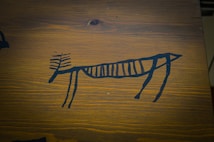 A simplistic, black drawing of an animal resembling a deer, created in a style reminiscent of ancient cave paintings, is placed against a wooden background with visible grain.