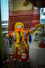 A collection of handmade dolls, dressed in colorful traditional garments and adorned with ribbons, displayed in front of a traditionally woven textile backdrop.
