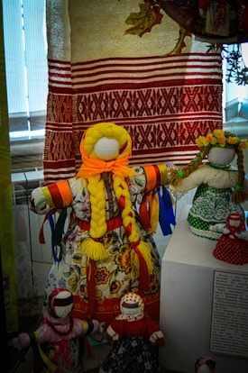 A collection of handmade dolls, dressed in colorful traditional garments and adorned with ribbons, displayed in front of a traditionally woven textile backdrop.