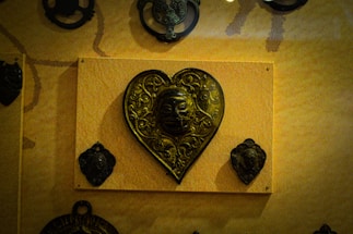 A heart-shaped metallic plaque with intricate carvings and a face in the center, mounted on a wooden board. Smaller metallic plaques with similar designs surround it.