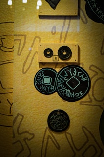 The image depicts an arrangement of various circular objects, likely ancient or historical coins, displayed on a yellowish patterned background. There is a wooden plaque with two circular items labeled '8' positioned in the upper center of the image. Below the plaque, there are multiple coins with inscriptions that appear to be in an ancient script.