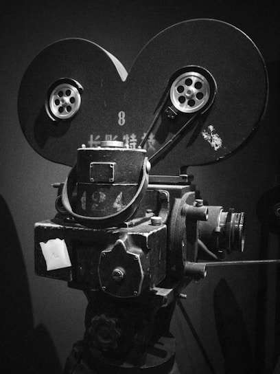A vintage film camera with large reels and intricate mechanical components. The camera has a classic design with visible spools and a lens. It appears to be made of metal with a dark finish, showcasing signs of wear.
