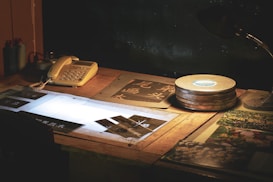 A wooden desk is illuminated by a dim light. On the desk, there is a vintage telephone, some film reels, and several photographic slides being viewed on a lightbox. There are also some printed photographs and other papers scattered around. The overall setting looks like an old-fashioned studio or a workspace.