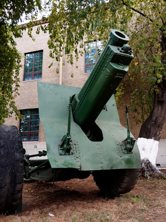 A large, green artillery gun is displayed outdoors on a patch of dirt and grass. It is positioned in front of a brick building with large windows. The scene is partially shaded by tall, leafy trees surrounding the gun.