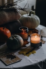 Various autumn-themed items, including small pumpkins, candles, and dried leaves, are arranged on a table. The scene has a cozy, warm atmosphere, with soft lighting and shadows. A stack of knitted blankets or sweaters is visible in the background, adding to the seasonal ambiance.