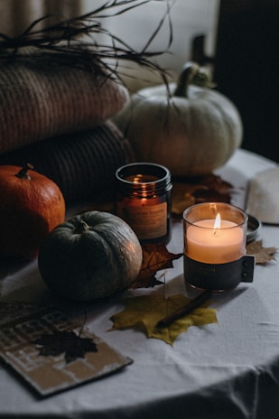 Various autumn-themed items, including small pumpkins, candles, and dried leaves, are arranged on a table. The scene has a cozy, warm atmosphere, with soft lighting and shadows. A stack of knitted blankets or sweaters is visible in the background, adding to the seasonal ambiance.