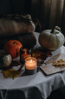 A cozy autumnal scene featuring a white tablecloth with various items arranged on it. There are small pumpkins, a lit candle in a glass jar, a closed jar, and several dried autumn leaves spread around. A couple of folded knit blankets are placed in the background, adding to the warm and rustic atmosphere.