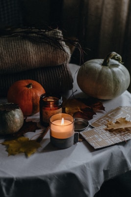 A cozy autumnal scene featuring a white tablecloth with various items arranged on it. There are small pumpkins, a lit candle in a glass jar, a closed jar, and several dried autumn leaves spread around. A couple of folded knit blankets are placed in the background, adding to the warm and rustic atmosphere.