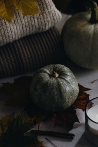 A small green pumpkin is surrounded by autumn leaves on a surface, alongside a folded knit sweater. There's a warm, cozy atmosphere with the use of soft textures and earthy tones.