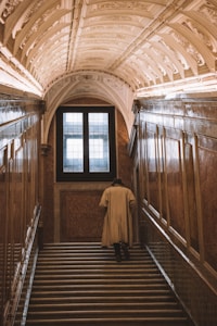 An elegantly dressed individual in a beige trench coat is walking up a grand staircase. The architecture is ornate, with an arched, decorated ceiling and richly paneled walls. A large, multi-pane window at the top of the stairs allows natural light to illuminate the space.