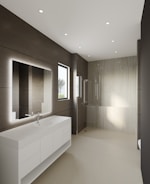 A modern bathroom features large, smooth tiles on the walls and floors, a sleek white countertop with built-in sink and storage cabinets, and a large illuminated mirror. Recessed ceiling lights provide ample illumination, and a walk-in shower with a glass partition is situated at the far end. A window allows natural light to enter, enhancing the minimalist aesthetic.