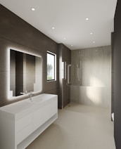 A modern bathroom features large, smooth tiles on the walls and floors, a sleek white countertop with built-in sink and storage cabinets, and a large illuminated mirror. Recessed ceiling lights provide ample illumination, and a walk-in shower with a glass partition is situated at the far end. A window allows natural light to enter, enhancing the minimalist aesthetic.