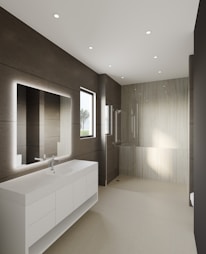 A modern bathroom features large, smooth tiles on the walls and floors, a sleek white countertop with built-in sink and storage cabinets, and a large illuminated mirror. Recessed ceiling lights provide ample illumination, and a walk-in shower with a glass partition is situated at the far end. A window allows natural light to enter, enhancing the minimalist aesthetic.