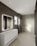 A modern bathroom features large, smooth tiles on the walls and floors, a sleek white countertop with built-in sink and storage cabinets, and a large illuminated mirror. Recessed ceiling lights provide ample illumination, and a walk-in shower with a glass partition is situated at the far end. A window allows natural light to enter, enhancing the minimalist aesthetic.
