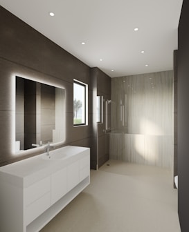 A modern bathroom features large, smooth tiles on the walls and floors, a sleek white countertop with built-in sink and storage cabinets, and a large illuminated mirror. Recessed ceiling lights provide ample illumination, and a walk-in shower with a glass partition is situated at the far end. A window allows natural light to enter, enhancing the minimalist aesthetic.