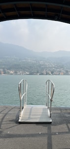 A metal platform with handrails extends over a calm body of water. In the background, there are distant hills and a shoreline dotted with buildings under a hazy sky.
