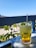 A refreshing beverage in a tall glass garnished with mint leaves and served with ice cubes sits on a table with a blue and white checkered tablecloth. In the background, there is a view of greenery and a path with people in a relaxed outdoor setting. The sky is clear and blue, suggesting a sunny day.
