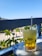 A refreshing beverage in a tall glass garnished with mint leaves and served with ice cubes sits on a table with a blue and white checkered tablecloth. In the background, there is a view of greenery and a path with people in a relaxed outdoor setting. The sky is clear and blue, suggesting a sunny day.