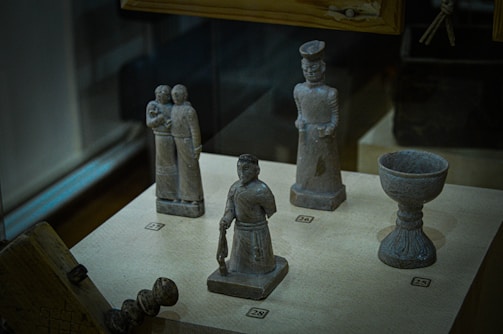 A display case contains several small, sculpted figurines and objects made of a stone-like material. There are figures dressed in traditional or historical attire, some holding objects, arranged neatly on a flat surface with numbered labels.