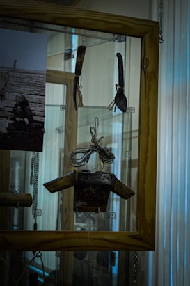 A display case contains various objects, including vintage tools and a photograph. The tools are arranged inside the glass with tags numbered from 9 to 12. The photograph, in black and white, is on the left side and features an outdoor scene with a person sitting on wooden planks. The background of the display case includes wooden elements and metal chains for support.