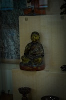 A statue of a seated figure, possibly a monk or religious figure, is displayed in a glass case. The figure is in a meditative position with one hand raised and appears to be wearing traditional robes. The statue has a dark, weathered appearance with hints of green and gold detailing. There are other artifacts, such as a small ornate cup, placed on adjacent shelves.