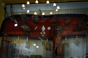 A display case contains a collection of tribal artifacts including a red fabric adorned with feathers, small metal objects in human and crescent moon shapes, and tassels hanging from the bottom edge. Reflections on the glass indicate overhead lighting, contributing to a museum-like setting.