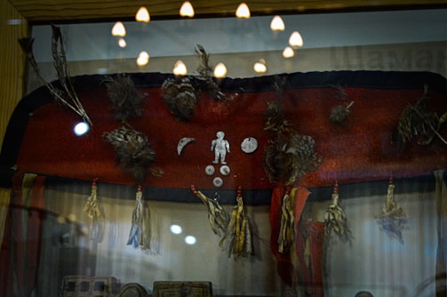 A display case contains a collection of tribal artifacts including a red fabric adorned with feathers, small metal objects in human and crescent moon shapes, and tassels hanging from the bottom edge. Reflections on the glass indicate overhead lighting, contributing to a museum-like setting.