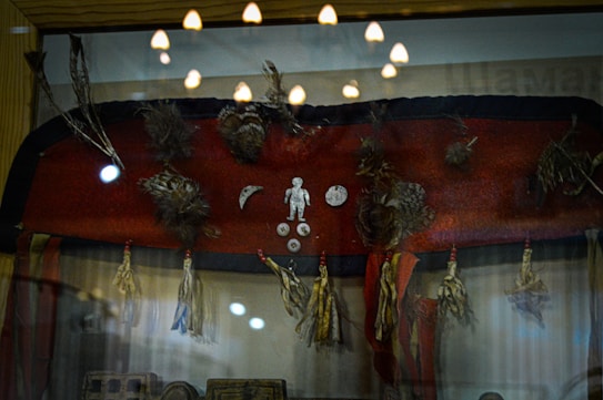 A display case contains a collection of tribal artifacts including a red fabric adorned with feathers, small metal objects in human and crescent moon shapes, and tassels hanging from the bottom edge. Reflections on the glass indicate overhead lighting, contributing to a museum-like setting.