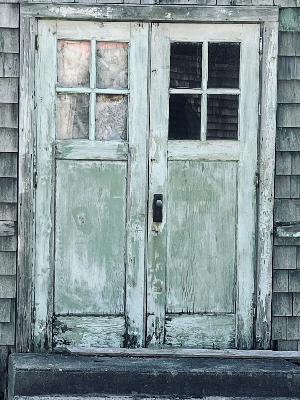 The image shows a weathered wooden door with two windows, one on each side and a small window at the top right corner. The door is painted green and has peeling paint around its edges, indicating it's an old building or structure. The door appears to be slightly ajar, suggesting it might have been left open for some time.