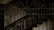 An intricate wrought iron fence features ornate swirling patterns and decorative elements with a staircase visible in the background, suggesting an entryway or barrier to a building. The lighting is dim, casting shadows on the details.