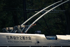 A close-up view of electric components and infrastructure, possibly on a railway vehicle or tram. Two large metallic rods with electrical insulators extend upward, connected to a frame attached to the vehicle. The surface has text in both Chinese and English, indicating 'World Design Cities Conference.' The background is dark with a blend of foliage and wires.
