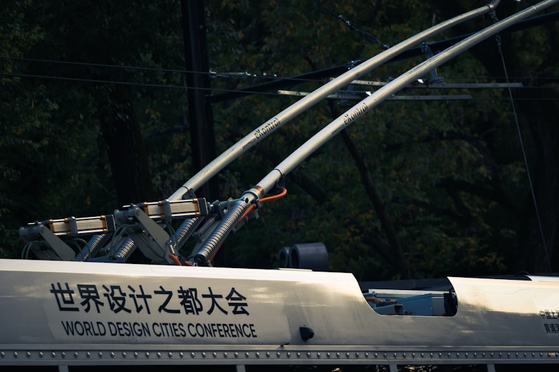 A close-up view of electric components and infrastructure, possibly on a railway vehicle or tram. Two large metallic rods with electrical insulators extend upward, connected to a frame attached to the vehicle. The surface has text in both Chinese and English, indicating 'World Design Cities Conference.' The background is dark with a blend of foliage and wires.