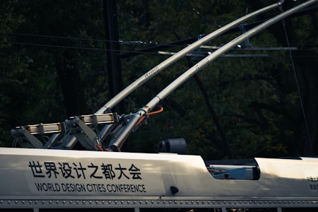A close-up view of electric components and infrastructure, possibly on a railway vehicle or tram. Two large metallic rods with electrical insulators extend upward, connected to a frame attached to the vehicle. The surface has text in both Chinese and English, indicating 'World Design Cities Conference.' The background is dark with a blend of foliage and wires.