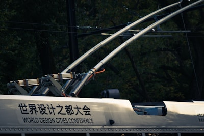 A close-up view of electric components and infrastructure, possibly on a railway vehicle or tram. Two large metallic rods with electrical insulators extend upward, connected to a frame attached to the vehicle. The surface has text in both Chinese and English, indicating 'World Design Cities Conference.' The background is dark with a blend of foliage and wires.