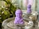 A small purple figurine of a meditating monk is placed on a glass surface filled with pebbles. In the background, another identical figurine is visible, enhancing the sense of calm. Surrounding the scene are blurred green leaves and a softly lit background, providing a serene setting.