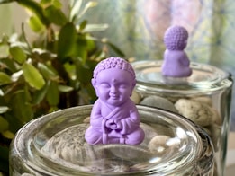 A small purple figurine of a meditating monk is placed on a glass surface filled with pebbles. In the background, another identical figurine is visible, enhancing the sense of calm. Surrounding the scene are blurred green leaves and a softly lit background, providing a serene setting.