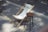 A white canvas deck chair is set up on a paved sidewalk next to a round wooden stool with black legs. The setting is outdoors on a sunny day, with patches of sunlight and shadows cast on the pavement. Yellow lines are painted on the ground, indicating parking or traffic regulation.