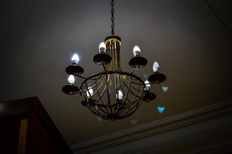 A large, ornate chandelier with a series of candle-like light bulbs arranged in a circular pattern is suspended from the ceiling. The lighting gives off a warm and dim ambiance, casting soft shadows and reflections on the ceiling and walls. The chandelier features intricate metalwork in a vintage style.